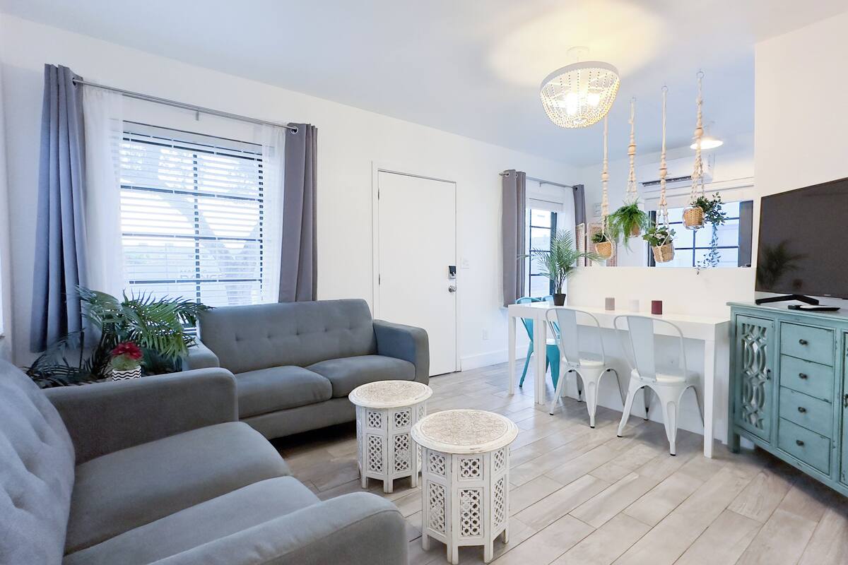 A modern living room with a grey sofa, two small side tables, a dining area with a white table and chairs, and a TV on a cabinet.