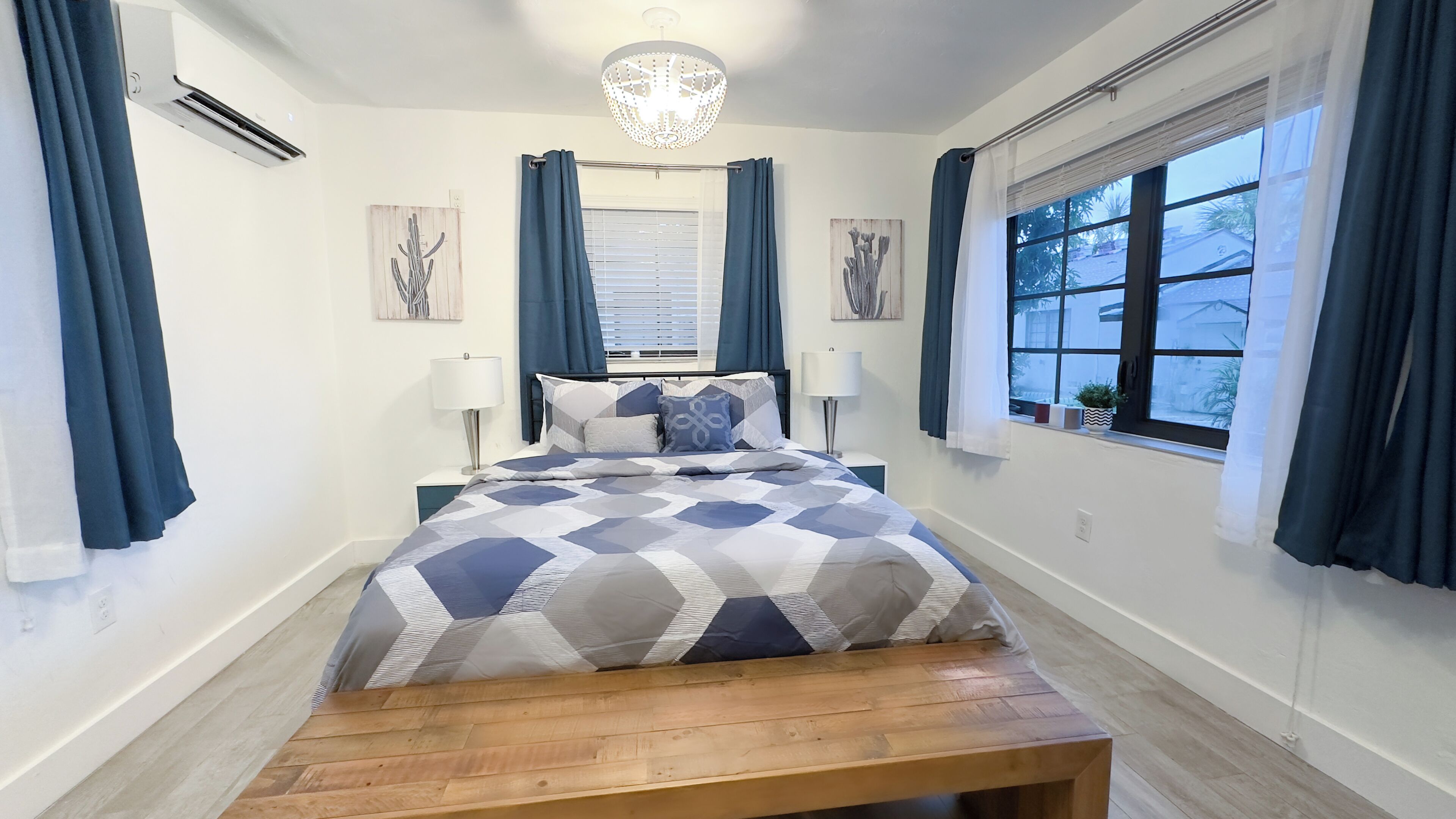 Um quarto com cama, um banquinho, um candelabro, janela com cortinas e obras de arte nas paredes.