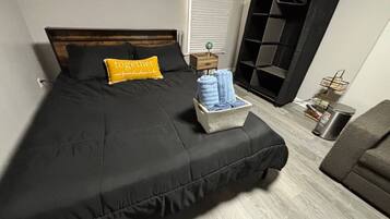 A neatly made bed with a black bedspread, a yellow pillow with text, and a wooden nightstand with a lamp.