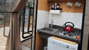 A compact kitchen with a sink, stove, and a small refrigerator. There are shelves with cups and bottles, and a window with curtains.
