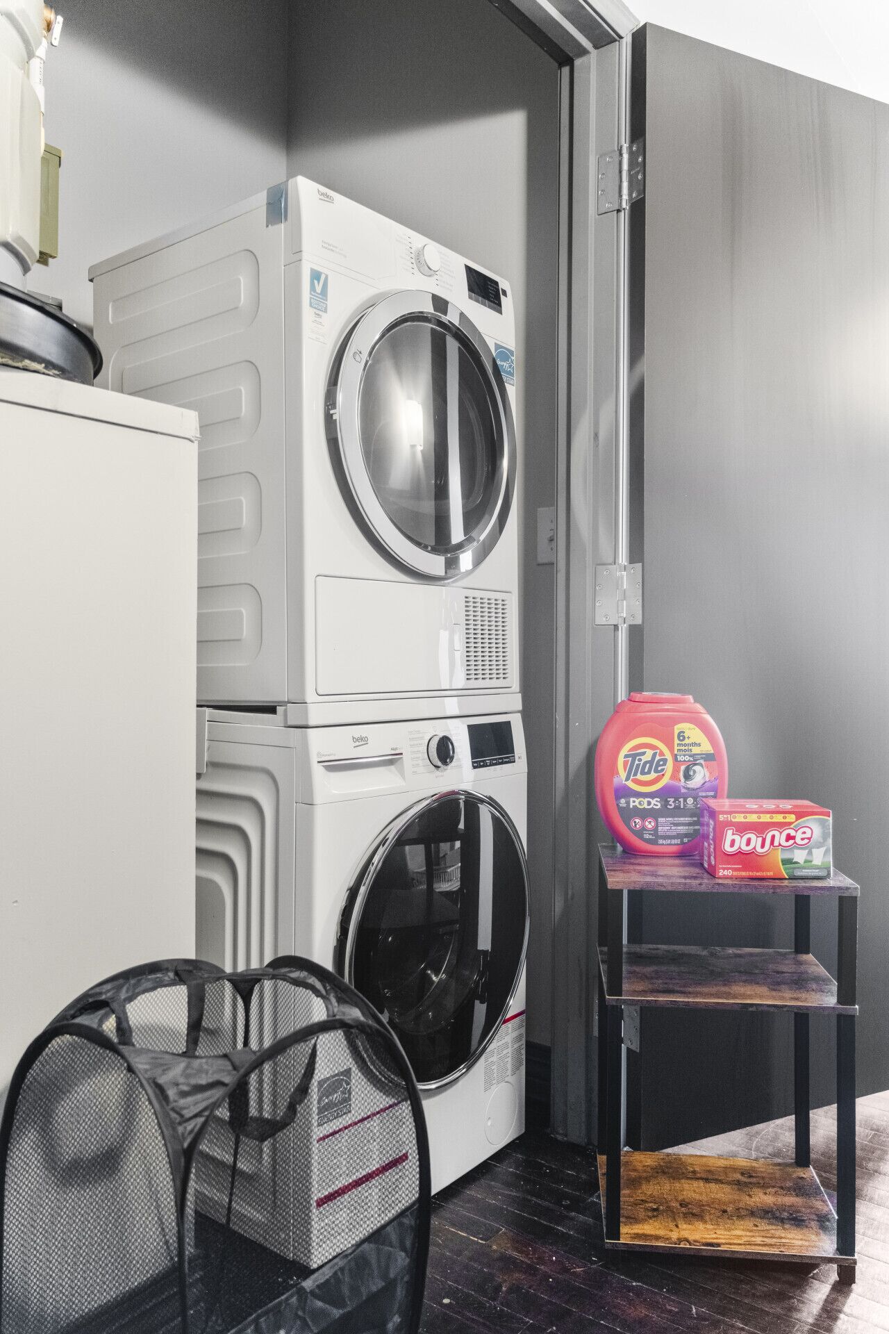 Un espace réduit dédié à la buanderie, comprenant un lave-linge et un sèche-linge à chargement frontal, une petite étagère avec de la lessive et un panier à linge en tissu à mailles.
