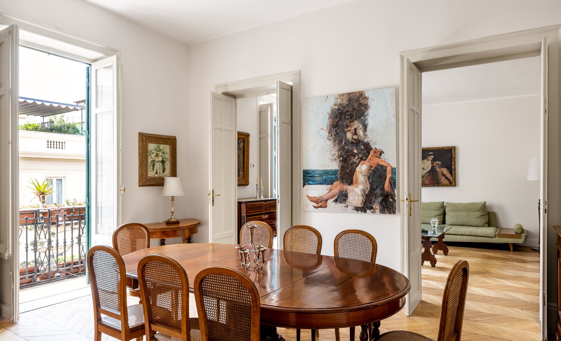 A dining area with a round wooden table and six wicker chairs. There is a painting on the wall and a green sofa in the background.
