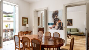 Een eethoek met een ronde houten tafel en zes rieten stoelen. Aan de muur hangt een schilderij en er staat een groene bank op de achtergrond.