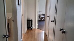 Un couloir avec un plancher en bois, des murs blancs et un luminaire au plafond. Il y a des portes sur le côté droit et un radiateur sur le côté gauche.