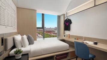A modern hotel room with a bed, a desk, a chair, a potted plant, and a city view.