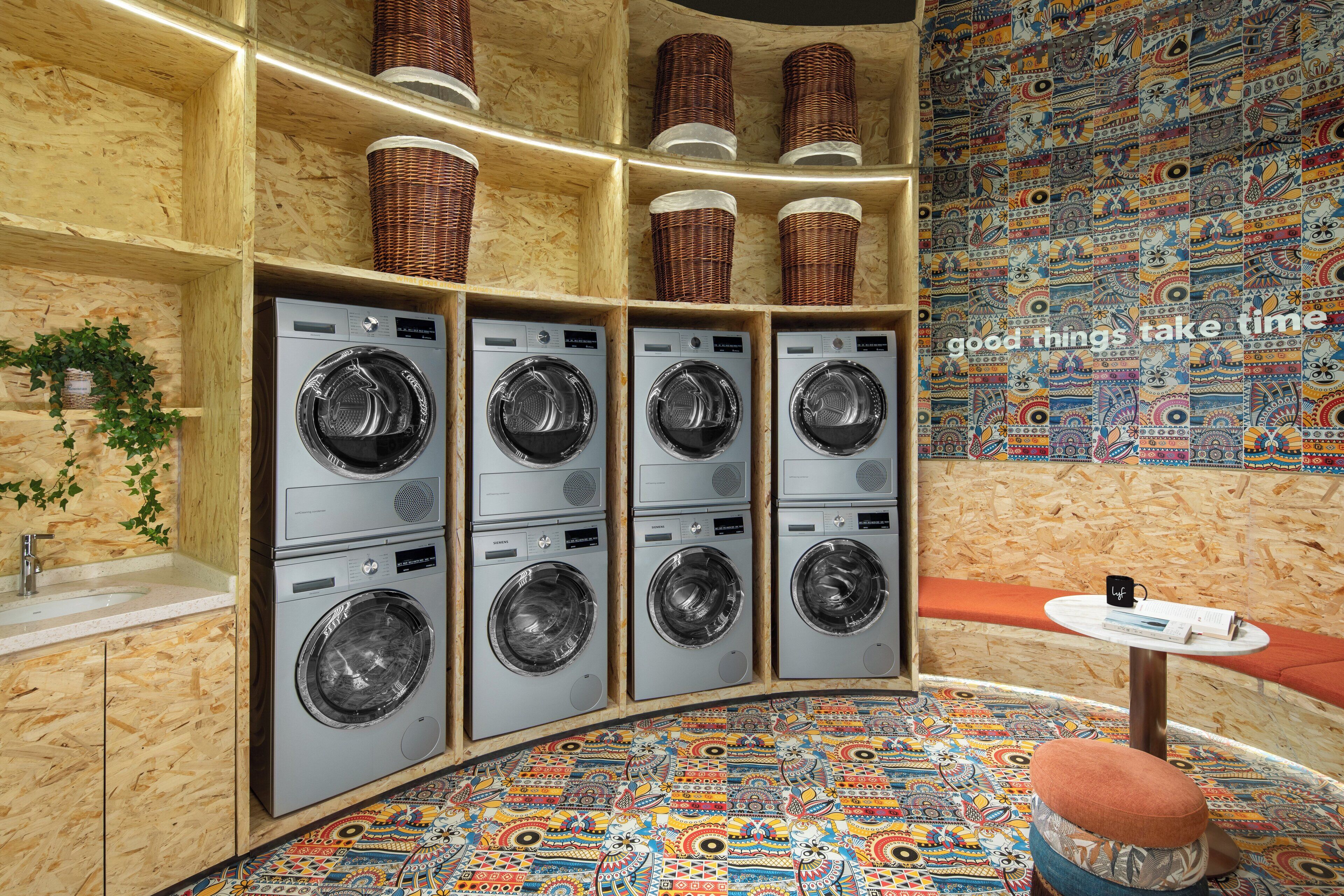 A laundry room with six washing machines, a sink, wicker baskets on shelves, a colorful patterned floor, and a small table with a mug and book.