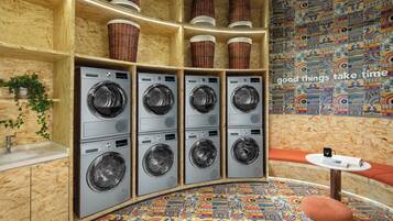 A laundry room with six washing machines, a sink, wicker baskets on shelves, a colorful patterned floor, and a small table with a mug and book.
