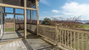 Une terrasse en bois avec une porte vitrée donnant sur un porche abrité, d’où l’on peut admirer une zone verdoyante avec des arbres et, au loin, une étendue d’eau.