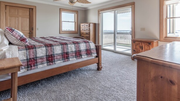 Une chambre à coucher comprenant un lit, une commode en bois, un ventilateur de plafond, une fenêtre et une porte-fenêtre coulissante donnant sur un balcon.