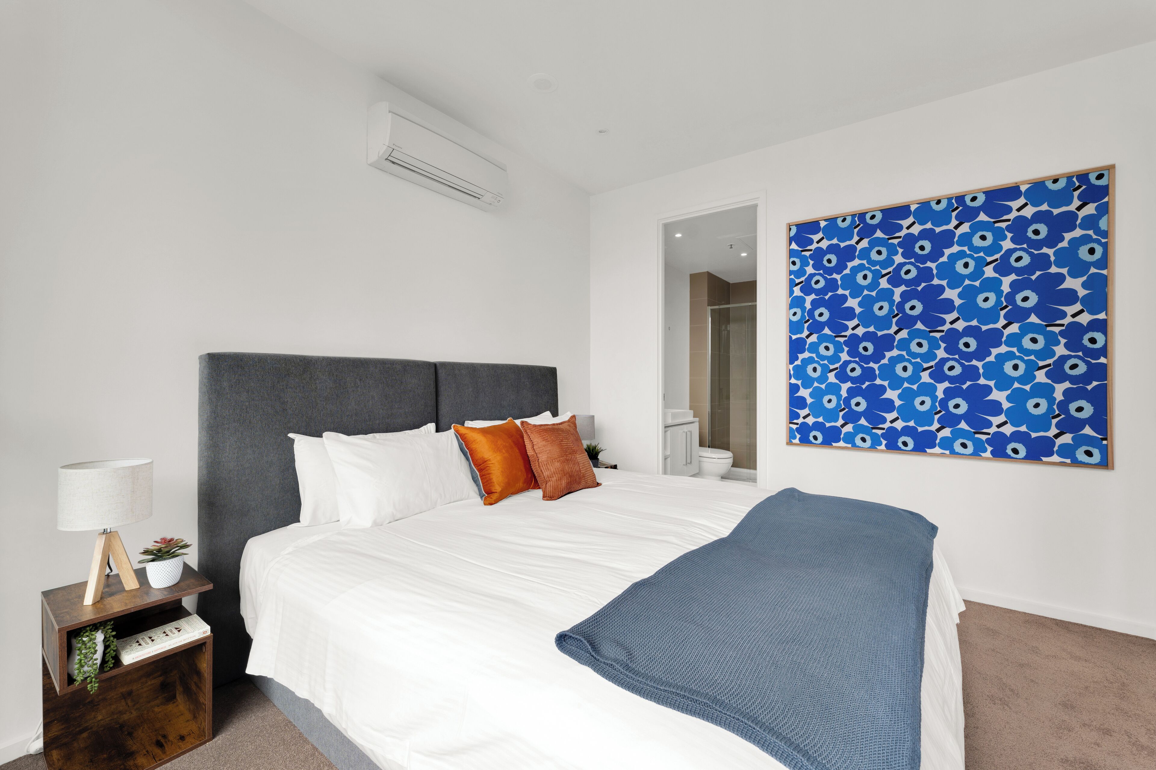 Ein Schlafzimmer mit einem Bett, einem Nachttisch, einer Lampe, einer blauen Decke und einem blauen floralen Kunstwerk an der Wand.