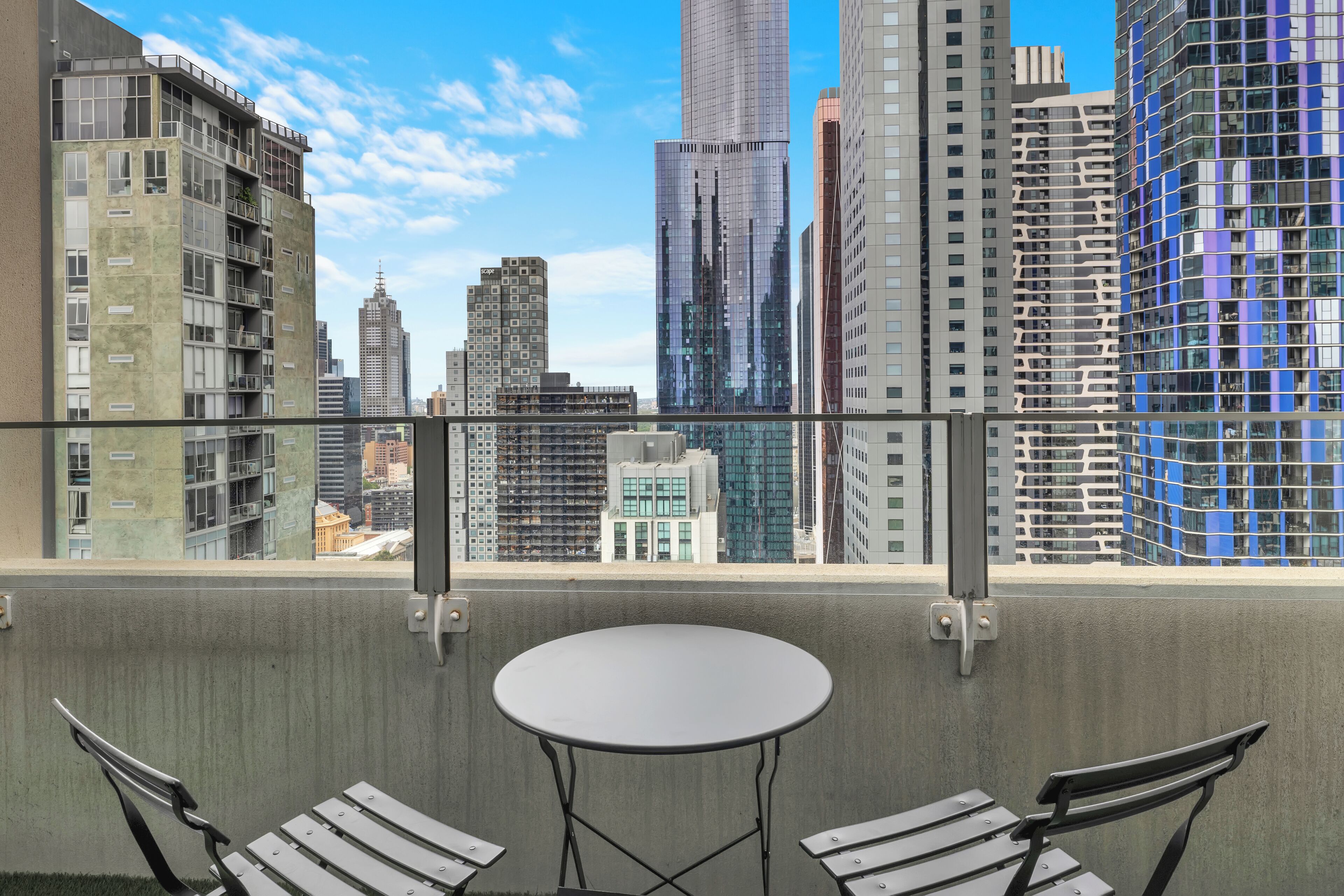 Un balcon avec une table ronde et deux chaises, donnant sur un paysage urbain avec des immeubles modernes de grande hauteur.