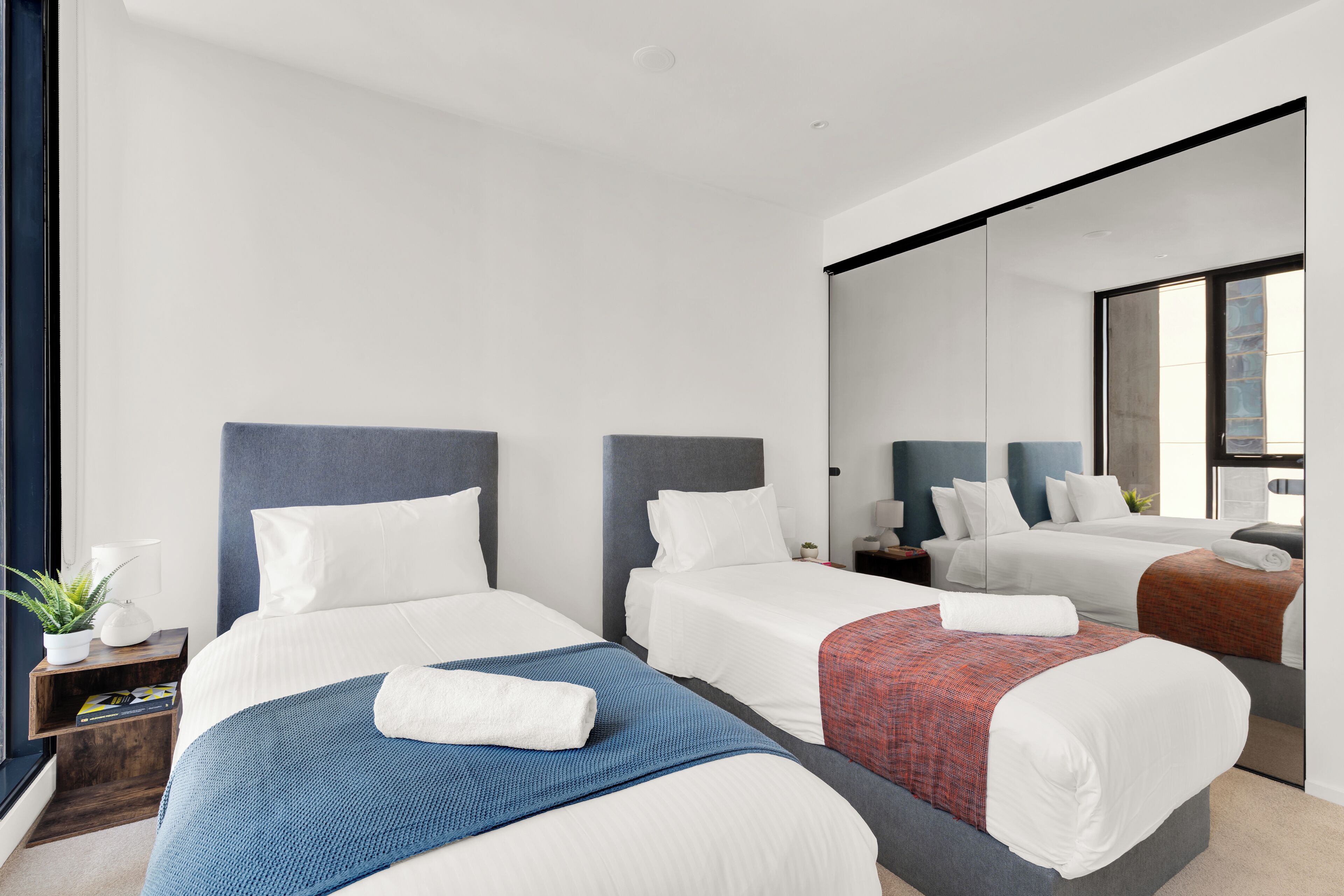 A hotel room with two beds, each with a headboard, white bedding, and a blue and red blanket. There is a small bedside table with a lamp and a plant.