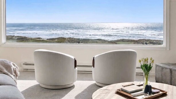 Een kamer met een groot raam dat uitkijkt op de oceaan, twee witte stoelen, een ronde tafel met een bloemenvaas, en een grijze bank.