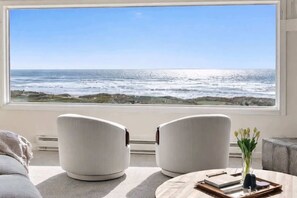 A room with a large window overlooking the ocean, two white chairs, a round table with a vase of flowers, and a gray sofa.