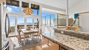 Een moderne keuken met een groot eiland, een eethoek met uitzicht op de oceaan, en een balkon met glazen wanden.