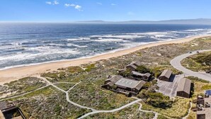 Luchtbeeld van een kustgebied met een strand, enkele gebouwen en een kronkelende weg.