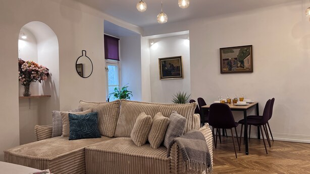 A modern living room with a sectional sofa, dining table, and chairs. There is a window with a view, a painting on the wall, and a plant.