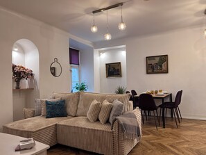 Una sala de estar moderna con un sofá seccional, una mesa de comedor y sillas. Se ve una ventana con vistas, un cuadro en la pared y una planta.