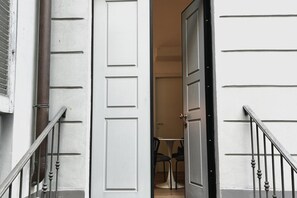 Un couloir avec une porte blanche, un escalier doté d’une rampe en métal et, au fond, une table entourée de chaises.