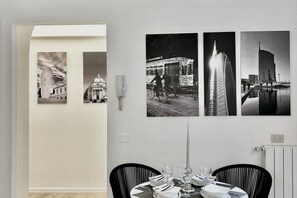 Een eetruimte met een tafel gedekt voor vier personen, drie zwart-wit foto's aan de muur, en een witte muur met een lichtschakelaar.