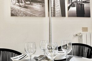 Uma mesa de jantar com três copos de vinho, um prato e um guardanapo. Há duas cadeiras com estofamento listrado em preto e branco. A parede apresenta duas fotografias em preto e branco.