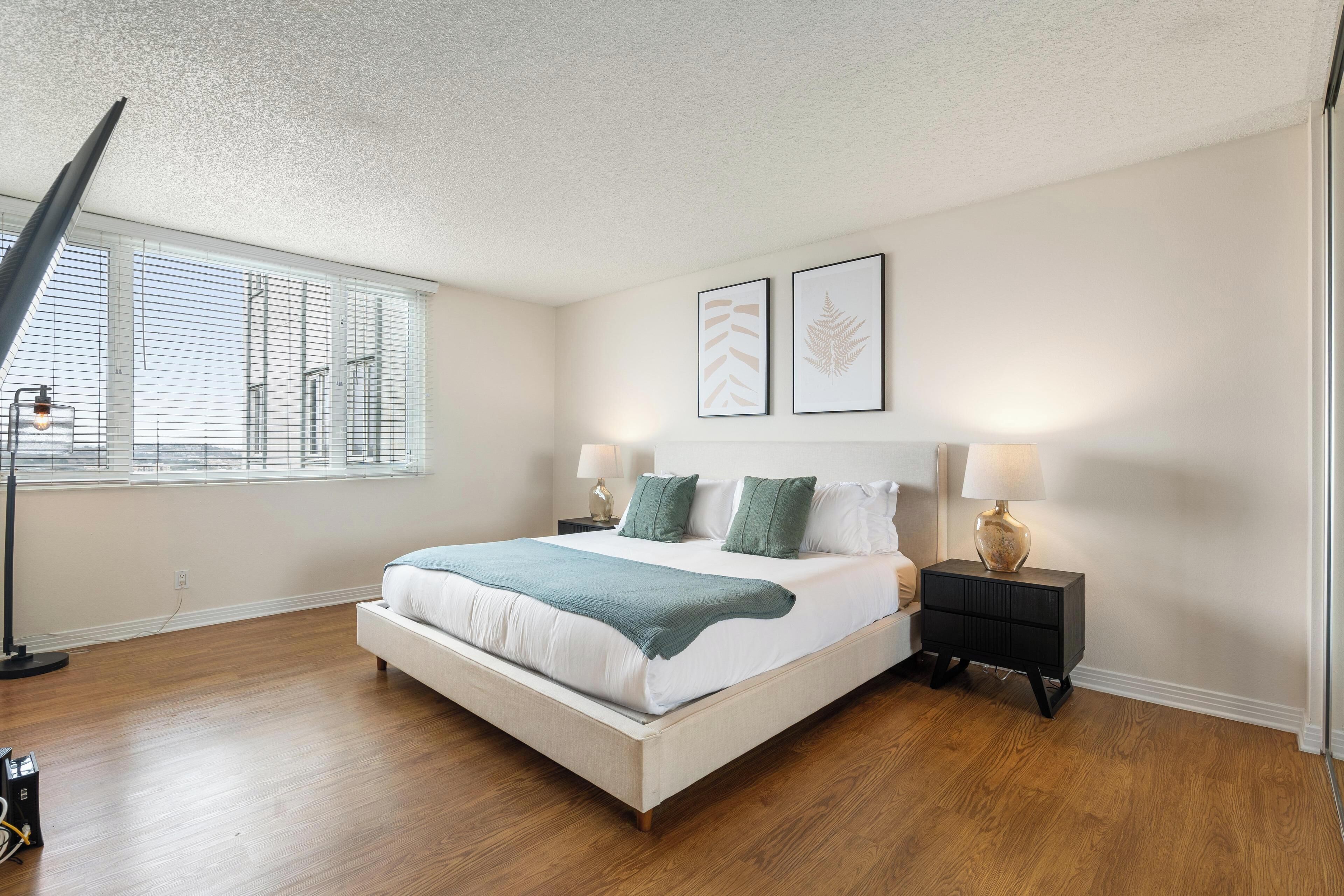 A bedroom with a large bed, two bedside tables, a floor lamp, and a window with blinds.