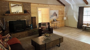 Una sala de estar acogedora con chimenea, paredes de madera, ventilador de techo y un gran ventanal con cortinas.