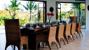 Een eetruimte met rieten stoelen, een lange tafel gedekt voor een maaltijd, en grote ramen met uitzicht op palmbomen en groen.