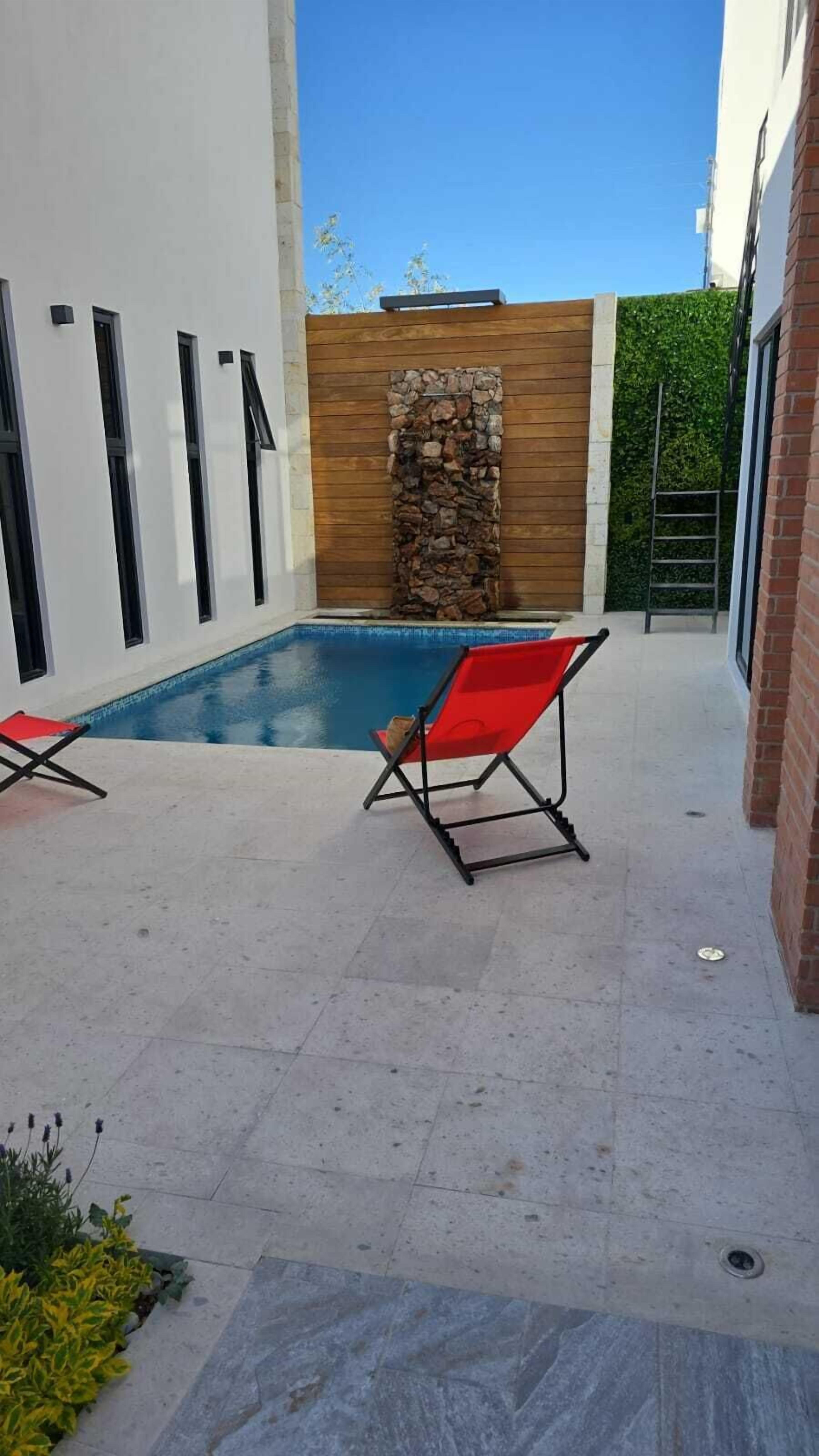 A modern outdoor pool area with two red lounge chairs, a stone wall, and a wooden fence.