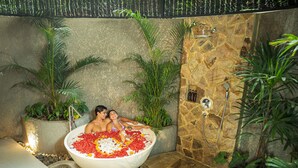 A couple relaxing in a large bathtub filled with flowers, surrounded by lush plants and a stone wall. - Emana Bambooscape (Ubud)