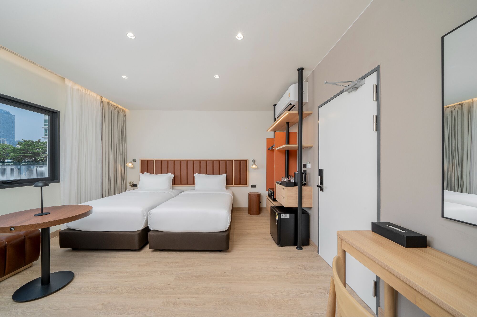 A hotel room with two beds, a desk, a chair, a small table, a trash bin, a mirror, and a window with curtains.