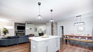 A modern kitchen with a central island, a dining area with a long table, and a living area with a sofa and TV.