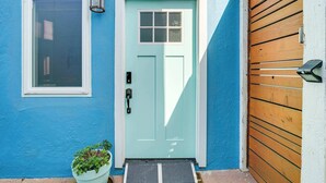 Une porte bleu clair avec une fenêtre au-dessus, un pot de fleurs placé à côté de la porte et une porte de garage en bois sur la droite.