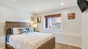 Une chambre à coucher avec un lit en bois, une table de chevet, une lampe, une fenêtre avec des stores et un téléviseur fixé au mur.