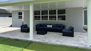 Un espacio de estar al aire libre techado con un sofá modular y dos sillones, una valla blanca y un cielo despejado.