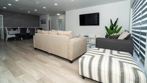 Una sala de estar moderna con un sofá color beige, un reposapiés a rayas y un sillón gris. Un televisor de pantalla plana está montado en la pared, y se observa una planta grande.