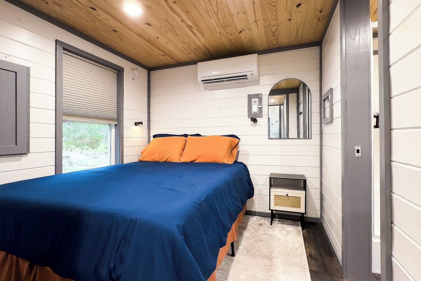 Un dormitorio con cama, aire acondicionado, mesita de noche, espejo y ventana con persianas.