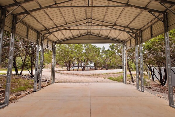 A covered pavilion with a concrete floor, metal beams, and a view of trees and a distant building.