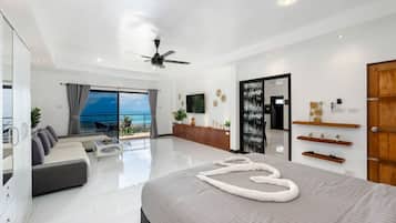 Une chambre d’hôtel moderne avec un grand lit, un balcon donnant sur la mer, un ventilateur de plafond et une porte en bois.