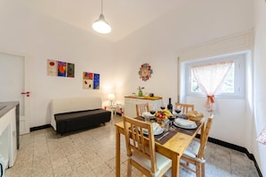 A dining area with a table set for two, a sofa, a small kitchen area, and a window with a curtain. - Graceful House in Monterosso, Monterosso, Italy (Monterosso)