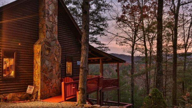 A log cabin with a stone chimney, a porch with red railings, and a view of the surrounding forest. - Rustic Cabin with Long Range Mountain Views - Hot Tub - Fire Pit (Blue Ridge)