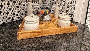Een keuken met een zwart-wit betegelde achterwand, een houten dienblad met twee witte theepotten en een roestvrijstalen waterkoker, en een aanrecht.