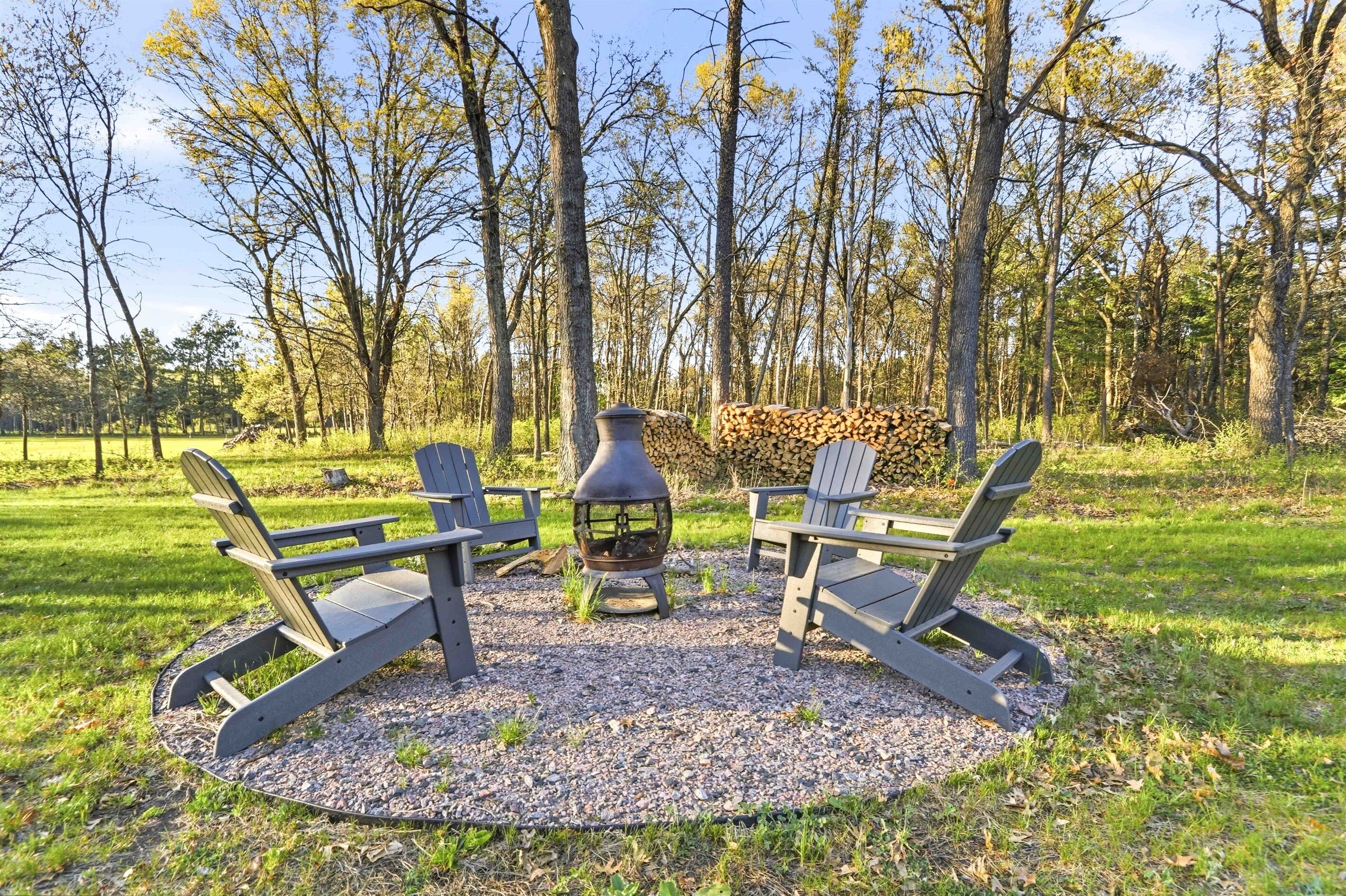 Outdoor firepit area with Adirondack seating, stacked firewood, and a peaceful wooded backdrop for relaxing evenings.