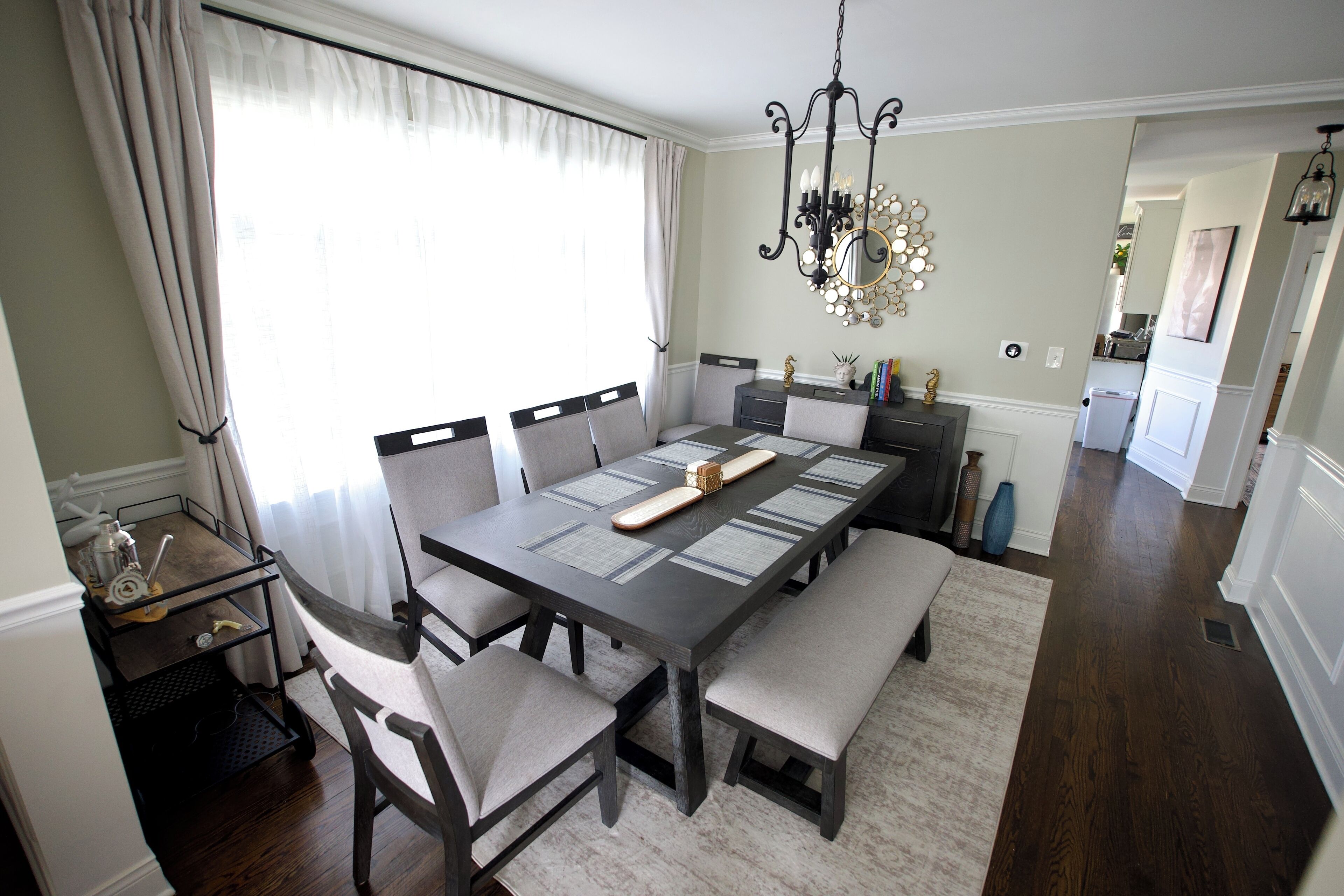 A dining area with a long table, multiple chairs, a chandelier, and a view into the kitchen.