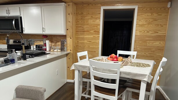 Una cocina compacta con armarios blancos, un microondas y una cafetera. Hay una mesa de comedor con cuatro sillas y un tazón de fruta. Se ve un banco al frente.