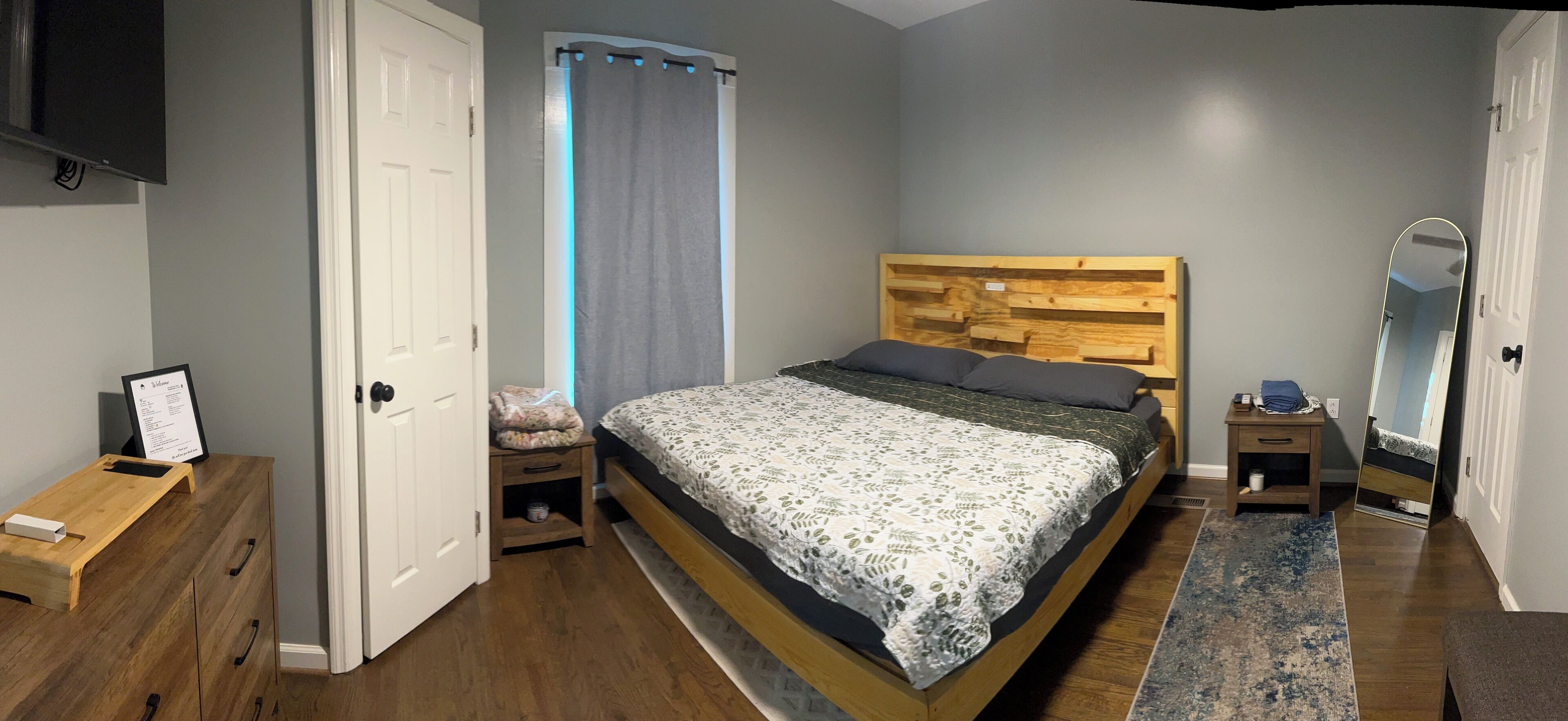 Un dormitorio con cabecera de madera, cama con colcha estampada, mesita de noche con lámpara, espejo y cómoda de madera.