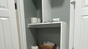 Un baño en esquina con puertas blancas, repisas con artículos de tocador, una cesta de mimbre y un perchero.