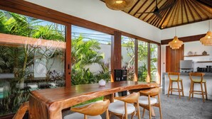 A modern kitchen with a wooden table and chairs, a coffee machine, and a view of lush greenery. - Casa Bojo & Casa Bella Uluwatu (Pecatu)