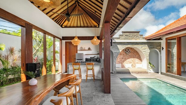 A modern outdoor dining area with a pool, wooden furniture, and a stone archway. - Casa Bojo & Casa Bella Uluwatu (Pecatu)