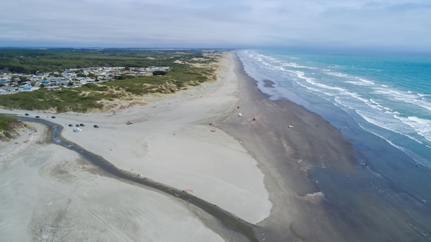 Una zona costera con una playa de arena, una carretera que serpentea por la orilla y un pequeño asentamiento con edificios al fondo.